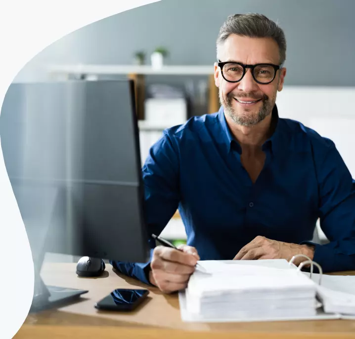 Um homem com camisa social azul sentado na sua cadeira sorrindo para tela do seu monitor.