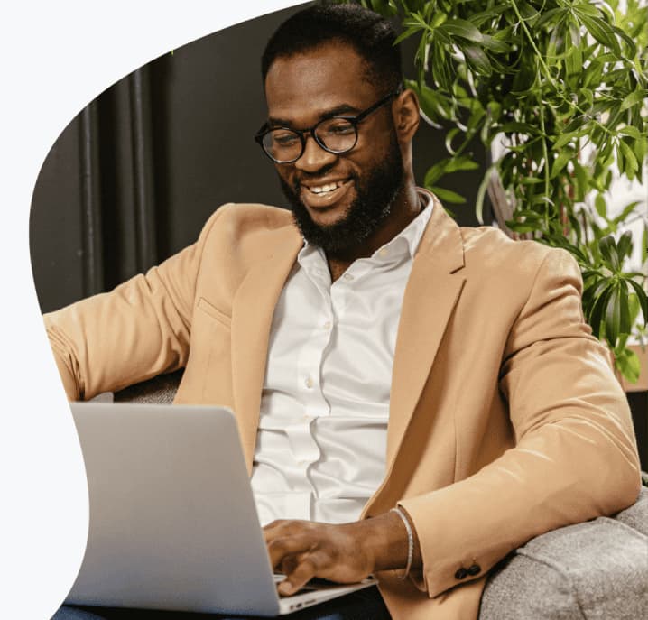 Um homem com terno cor de pele sentado na poltrona sorrindo para tela do seu notebook.